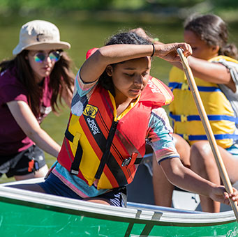 A photograph of two young girls paddeling a canoe with an instructor in the background helping.